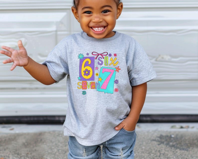May include: A gray t-shirt with the numbers "6" and "7" in colorful designs. The word "seven" is below the number "6". The shirt is worn by a young child.