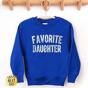 May include: A royal blue sweatshirt with the words "FAVORITE DAUGHTER" printed in white, block letters. The sweatshirt has long sleeves and a crew neck. The garment is hanging on a wooden hanger.