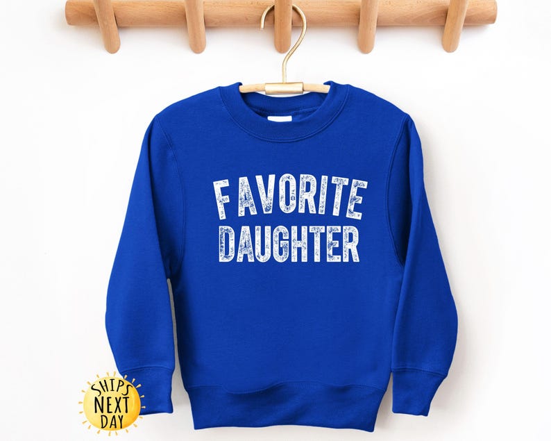May include: A royal blue sweatshirt with the words "FAVORITE DAUGHTER" printed in white, block letters. The sweatshirt has long sleeves and a crew neck. The garment is hanging on a wooden hanger.