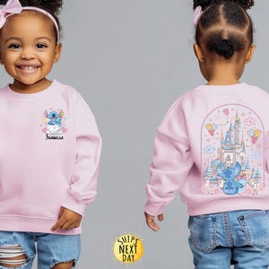 May include: A light pink sweatshirt featuring a cartoon Stitch design. The front shows Stitch in a teacup with the name "Isabella". The back displays a castle scene with Stitch. The sweatshirt is worn by a young child.