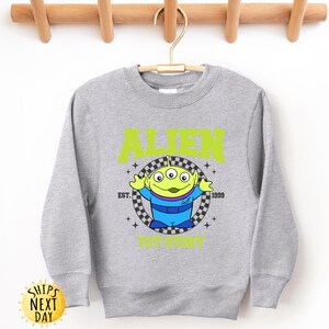 Puede incluir: Una sudadera gris con un personaje extraterrestre verde de Toy Story. El texto "Alien" est&aacute; impreso en amarillo sobre el personaje y "Est. 1999" est&aacute; impreso en amarillo debajo del personaje. El texto "Toy Story" est&aacute; impreso en amarillo debajo del personaje.