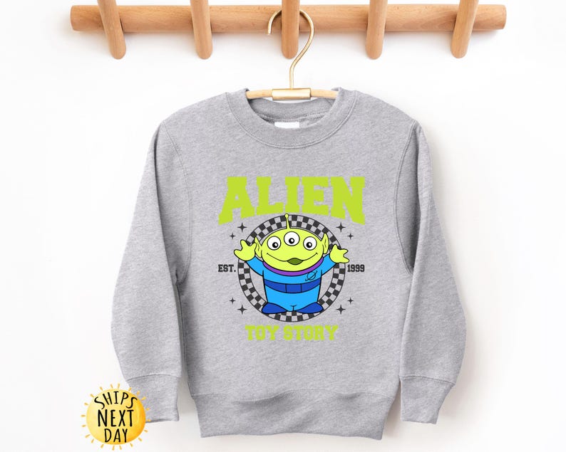 Puede incluir: Una sudadera gris con un personaje extraterrestre verde de Toy Story. El texto "Alien" est&aacute; impreso en amarillo sobre el personaje y "Est. 1999" est&aacute; impreso en amarillo debajo del personaje. El texto "Toy Story" est&aacute; impreso en amarillo debajo del personaje.