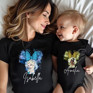 May include: A black short-sleeved t-shirt featuring a blue watercolor Minnie Mouse ear design with Elsa and the name Isabella, and a black short-sleeved baby bodysuit featuring a green watercolor Minnie Mouse ear design with Tiana and the name Amelia.