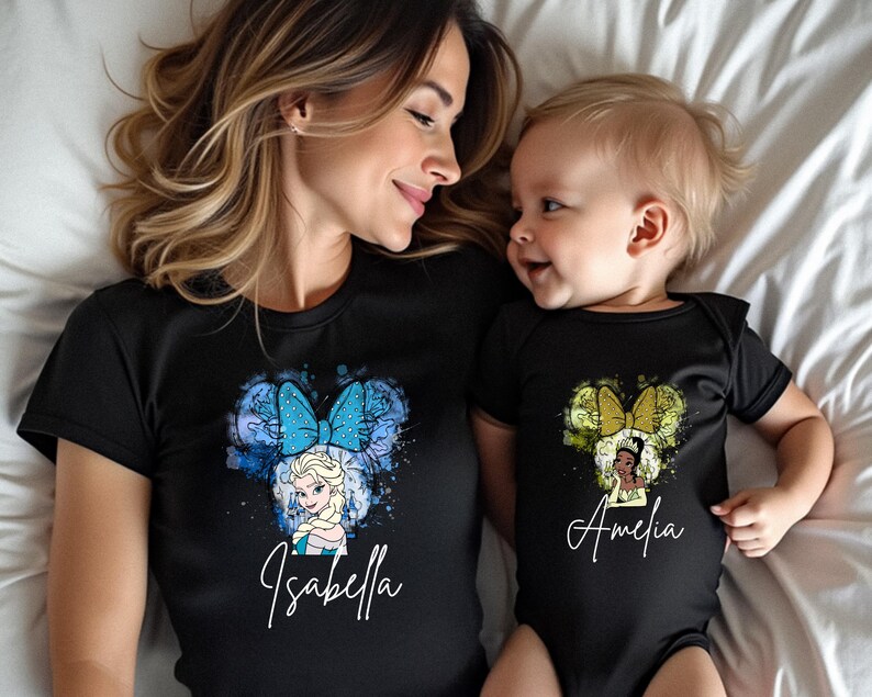 May include: A black short-sleeved t-shirt featuring a blue watercolor Minnie Mouse ear design with Elsa and the name Isabella, and a black short-sleeved baby bodysuit featuring a green watercolor Minnie Mouse ear design with Tiana and the name Amelia.