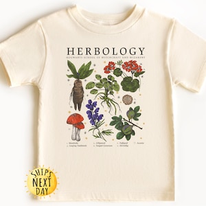 May include: A beige t-shirt with a graphic print of various plants and mushrooms. The text "HERBOLOGY" is printed at the top of the graphic. The text "HOGWARTS SCHOOL OF WITCHCRAFT AND WIZARDRY" is printed below the word "HERBOLOGY".