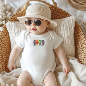 May include: A baby wearing a white bodysuit with a colorful graphic of superheroes. The baby is also wearing a straw hat and sunglasses, and is sitting in a woven basket.