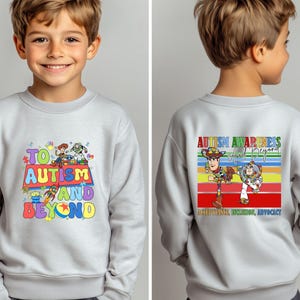 K&ouml;nnte beinhalten: Ein hellgraues Sweatshirt mit dem Schriftzug "TO AUTISM AND BEYOND" und bunten Zeichentrickfiguren. Auf der R&uuml;ckseite des Sweatshirts steht "AUTISM AWARENESS And Beyond" mit Zeichentrickfiguren und einem Regenbogenstreifen-Design.