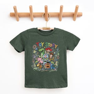 May include: A dark green short-sleeve t-shirt featuring a colorful graphic print of Toy Story characters including Woody, Buzz Lightyear, Rex, and Mr. Potato Head, surrounded by scribbled crayon-style drawings and the text TOY STORY.