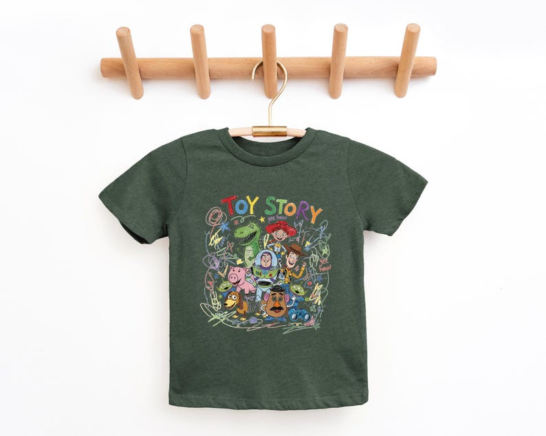 May include: A dark green short-sleeve t-shirt featuring a colorful graphic print of Toy Story characters including Woody, Buzz Lightyear, Rex, and Mr. Potato Head, surrounded by scribbled crayon-style drawings and the text TOY STORY.