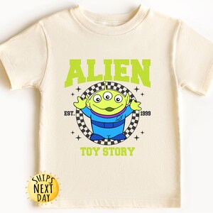 Puede incluir: Una camiseta verde claro con un personaje extraterrestre verde de Toy Story. La camiseta tiene el texto "ALIEN" en letras verdes sobre el extraterrestre y "EST. 1999" y "TOY STORY" debajo del extraterrestre.