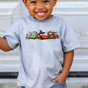 K&ouml;nnte beinhalten: Hellgraues T-Shirt mit einem Cartoon-Motiv von Autos zum Thema Halloween. Die Autos sind rot, gr&uuml;n und braun. Das T-Shirt wird von einem kleinen Kind getragen.