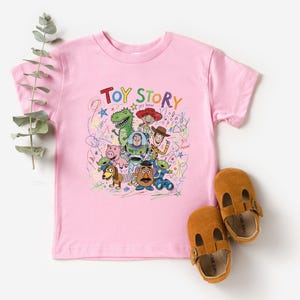 May include: A light pink short-sleeve t-shirt featuring a colorful graphic print of Toy Story characters including Woody, Buzz Lightyear, Jessie, Rex, and Mr. Potato Head, surrounded by scribbled drawings and the text Toy Story in multi-colored letters.