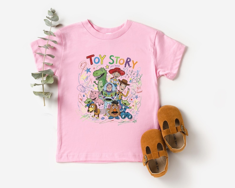 May include: A light pink short-sleeve t-shirt featuring a colorful graphic print of Toy Story characters including Woody, Buzz Lightyear, Jessie, Rex, and Mr. Potato Head, surrounded by scribbled drawings and the text Toy Story in multi-colored letters.