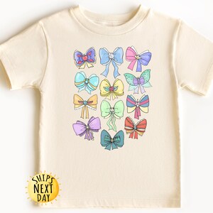 May include: A cream-colored t-shirt with a colorful design of bows. The bows are in various colors, including pink, blue, yellow, green, and purple.