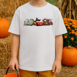 K&ouml;nnte beinhalten: Wei&szlig;es Kurzarm-T-Shirt mit einem farbenfrohen Aufdruck von Cartoon-Autos in Halloween-Kost&uuml;men. Das Design umfasst ein gr&uuml;nes Auto, ein rotes Auto mit Hexenhut und andere Fahrzeuge. Das T-Shirt wird von einem Kind getragen, mit K&uuml;rbissen und Herbstlaub im Hintergrund.