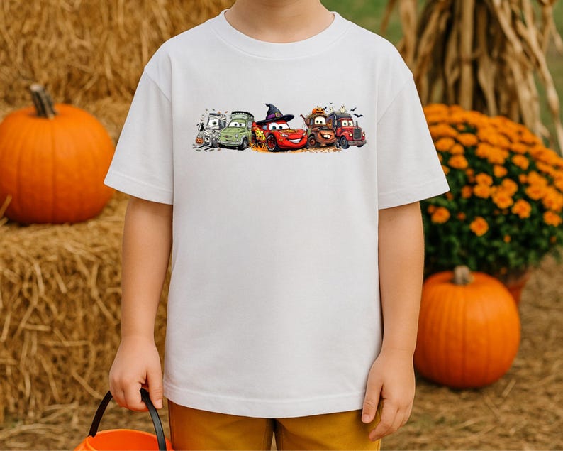 K&ouml;nnte beinhalten: Wei&szlig;es Kurzarm-T-Shirt mit einem farbenfrohen Aufdruck von Cartoon-Autos in Halloween-Kost&uuml;men. Das Design umfasst ein gr&uuml;nes Auto, ein rotes Auto mit Hexenhut und andere Fahrzeuge. Das T-Shirt wird von einem Kind getragen, mit K&uuml;rbissen und Herbstlaub im Hintergrund.