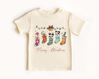 May include: A cream-coloured t-shirt with a Christmas-themed design. It features Disney characters in stockings, fairy lights, and the script text "Merry Christmas". A festive tee for the holiday season.