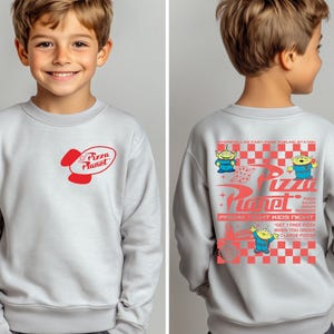Puede incluir: Sudadera gris claro con el logo de Pizza Planet en rojo en la parte delantera. La parte trasera de la sudadera muestra el logo de Pizza Planet, personajes alien&iacute;genas y texto en rojo, con un patr&oacute;n de cuadros rojos y blancos.