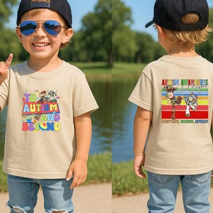 K&ouml;nnte beinhalten: Beiges kurz&auml;rmeliges T-Shirt mit einem farbenfrohen Aufdruck auf der Vorderseite mit dem Text "To Autism and Beyond" und Zeichentrickfiguren. Auf der R&uuml;ckseite des Shirts befindet sich ein Aufdruck mit der Aufschrift "Autism Awareness and Beyond".