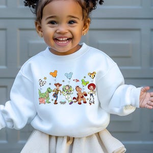 May include: White crewneck sweatshirt featuring a colorful graphic of Toy Story characters. The design includes Woody, Buzz Lightyear, Jessie, and other characters with balloon accents. A fun and playful design for kids.