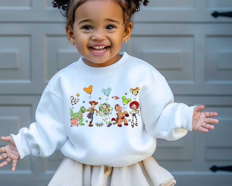 May include: White crewneck sweatshirt featuring a colorful graphic of Toy Story characters. The design includes Woody, Buzz Lightyear, Jessie, and other characters with balloon accents. A fun and playful design for kids.