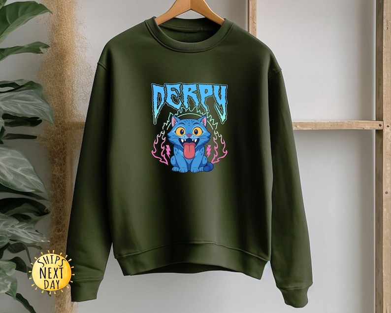 May include: An olive green sweatshirt with a cartoon cat design. The cat is blue with yellow eyes and a pink tongue. The word "DERPY" is above the cat in blue and white lettering. The cat is surrounded by pink and purple flames.