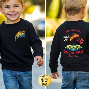 May include: Black sweatshirt featuring a colorful graphic of the Pizza Planet aliens and pizza slice. The back of the sweatshirt has the text "KEEP YOUR EYES ON THE PRIZE" and more graphics. The sweatshirt is worn by a child.