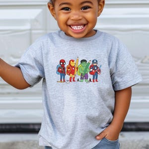 May include: A gray t-shirt featuring a colorful graphic of cartoon superheroes, including Spider-Man, Iron Man, Hulk, and Captain America, each holding school supplies. The design is printed on a short-sleeved shirt.
