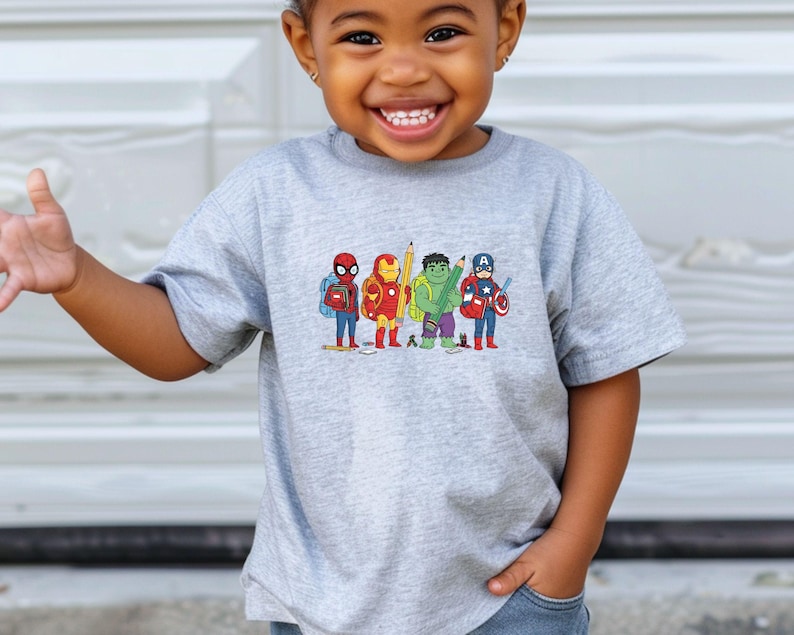 May include: A gray t-shirt featuring a colorful graphic of cartoon superheroes, including Spider-Man, Iron Man, Hulk, and Captain America, each holding school supplies. The design is printed on a short-sleeved shirt.