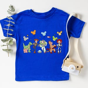 May include: A royal blue t-shirt featuring a colorful graphic of characters from the Toy Story movie. The design includes Woody, Buzz Lightyear, Jessie, and other characters. The shirt is a short-sleeved crew neck.