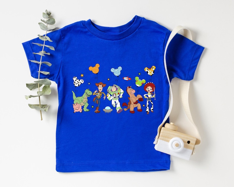 May include: A royal blue t-shirt featuring a colorful graphic of characters from the Toy Story movie. The design includes Woody, Buzz Lightyear, Jessie, and other characters. The shirt is a short-sleeved crew neck.