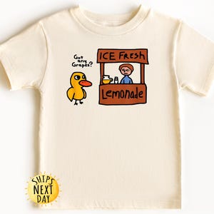 May include: Cream-colored t-shirt featuring a cartoon duck asking "Got any Grapes?" next to a lemonade stand that says "ICE FRESH Lemonade." The shirt has a round neck and short sleeves. A yellow sun graphic with the text "SHIPS NEXT DAY" is at the bottom.
