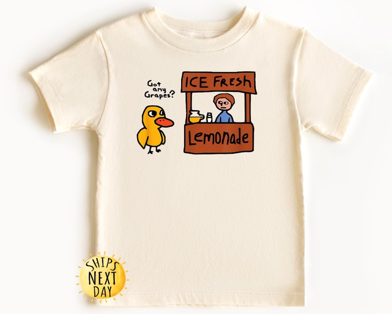 May include: Cream-colored t-shirt featuring a cartoon duck asking "Got any Grapes?" next to a lemonade stand that says "ICE FRESH Lemonade." The shirt has a round neck and short sleeves. A yellow sun graphic with the text "SHIPS NEXT DAY" is at the bottom.