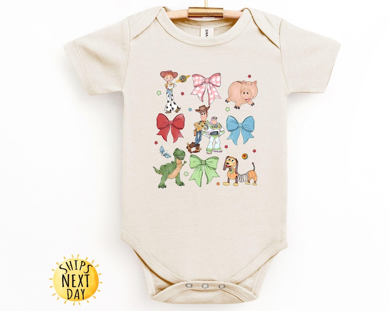 May include: A beige baby onesie with a print of Toy Story characters, including Woody, Buzz Lightyear, Rex, and Slinky Dog. The characters are surrounded by colorful bows.