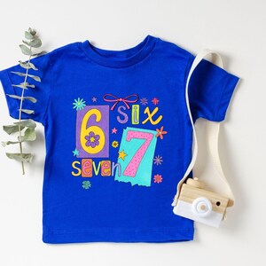May include: A royal blue t-shirt with the numbers "6" and "7" in colorful designs. The word "Six" and "Seven" are also displayed. A small wooden camera with a white strap is on the right side of the shirt.