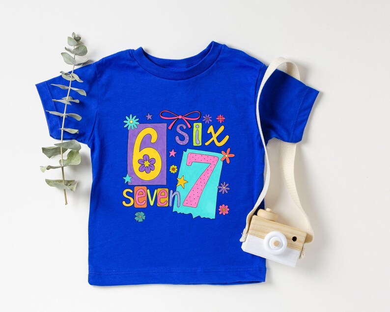 May include: A royal blue t-shirt with the numbers "6" and "7" in colorful designs. The word "Six" and "Seven" are also displayed. A small wooden camera with a white strap is on the right side of the shirt.