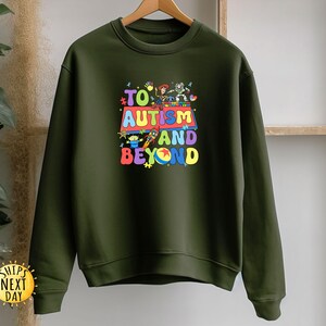 K&ouml;nnte beinhalten: Ein olivgr&uuml;nes Sweatshirt mit einem farbenfrohen Aufdruck mit der Aufschrift "TO AUTISM AND BEYOND". Die Grafik zeigt Zeichentrickfiguren und ein mehrfarbiges Design. Das Sweatshirt h&auml;ngt an einem Holzb&uuml;gel.
