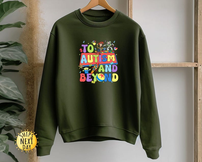 K&ouml;nnte beinhalten: Ein olivgr&uuml;nes Sweatshirt mit einem farbenfrohen Aufdruck mit der Aufschrift "TO AUTISM AND BEYOND". Die Grafik zeigt Zeichentrickfiguren und ein mehrfarbiges Design. Das Sweatshirt h&auml;ngt an einem Holzb&uuml;gel.