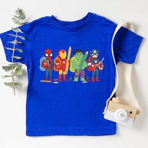 May include: A royal blue t-shirt featuring a graphic of four superheroes: Spider-Man, Iron Man, Hulk, and Captain America, each holding a pencil. The superheroes are depicted with backpacks, suggesting a back-to-school theme. A white strap with a wooden camera is on the right.