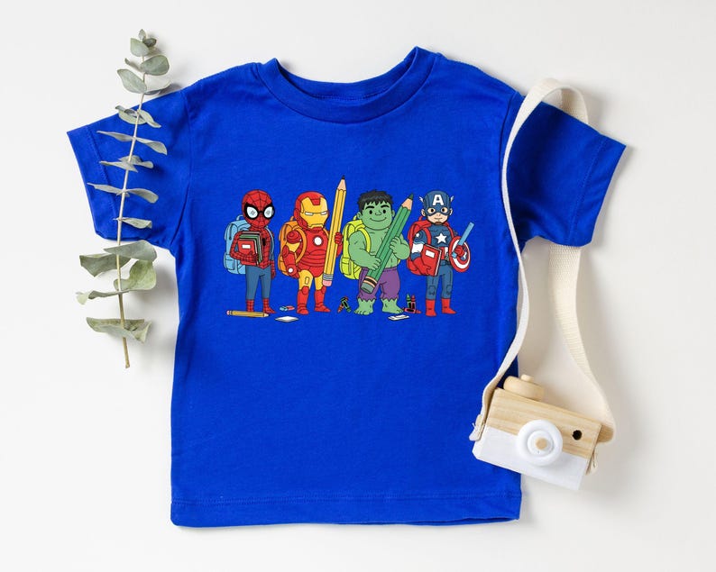 May include: A royal blue t-shirt featuring a graphic of four superheroes: Spider-Man, Iron Man, Hulk, and Captain America, each holding a pencil. The superheroes are depicted with backpacks, suggesting a back-to-school theme. A white strap with a wooden camera is on the right.