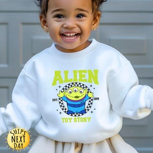 Puede incluir: Sudadera blanca con un personaje extraterrestre verde de Toy Story. El texto "Alien" est&aacute; en un cuadro amarillo a cuadros con el texto "Toy Story" debajo. El texto "Est. 1999" est&aacute; en un cuadro amarillo m&aacute;s peque&ntilde;o encima del extraterrestre.