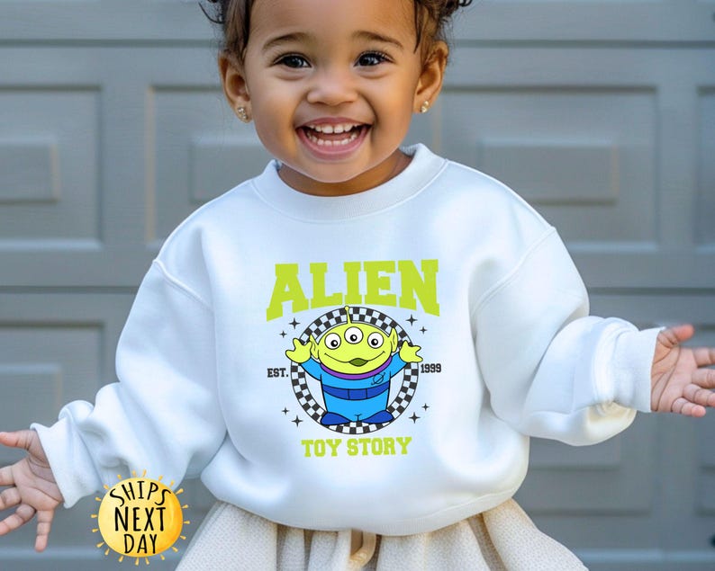 Puede incluir: Sudadera blanca con un personaje extraterrestre verde de Toy Story. El texto "Alien" est&aacute; en un cuadro amarillo a cuadros con el texto "Toy Story" debajo. El texto "Est. 1999" est&aacute; en un cuadro amarillo m&aacute;s peque&ntilde;o encima del extraterrestre.
