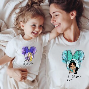 May include: Two white t-shirts featuring graphic prints of cartoon characters with mouse ears. The smaller shirt displays a blonde character with the name Olivia, while the larger shirt features a dark-haired character with the name Liam.