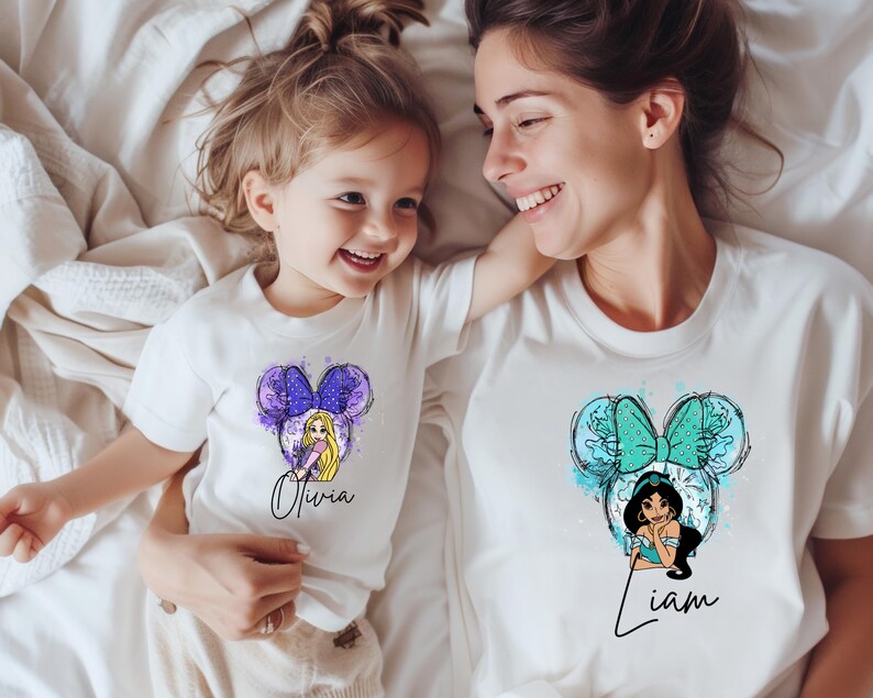 May include: Two white t-shirts featuring graphic prints of cartoon characters with mouse ears. The smaller shirt displays a blonde character with the name Olivia, while the larger shirt features a dark-haired character with the name Liam.