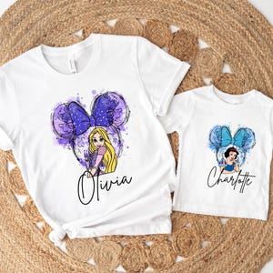 May include: Two white t-shirts featuring graphic prints. The larger shirt displays a Rapunzel character with a purple bow and mouse ears, with the name Olivia below. The smaller shirt shows a Snow White character with a blue bow and mouse ears, with the name Charlotte below.