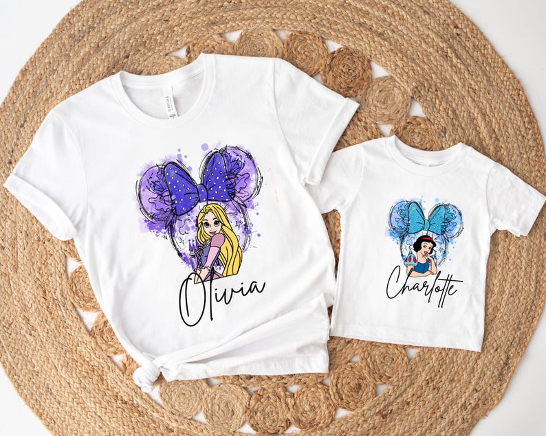 May include: Two white t-shirts featuring graphic prints. The larger shirt displays a Rapunzel character with a purple bow and mouse ears, with the name Olivia below. The smaller shirt shows a Snow White character with a blue bow and mouse ears, with the name Charlotte below.