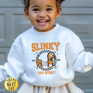 May include: White sweatshirt with a graphic of Slinky Dog from Toy Story. The graphic includes the text "Slinky" in a black and orange checkered design, "Est. 1995", and "Toy Story".