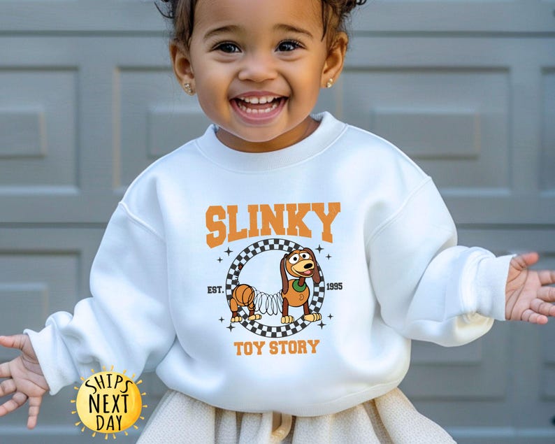 May include: White sweatshirt with a graphic of Slinky Dog from Toy Story. The graphic includes the text "Slinky" in a black and orange checkered design, "Est. 1995", and "Toy Story".