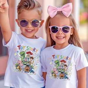 May include: Two white short-sleeved t-shirts featuring a colorful graphic print of Toy Story characters including Woody, Buzz Lightyear, Rex, and Mr. Potato Head, with the text Toy Story printed in a multi-colored font above the character illustration.