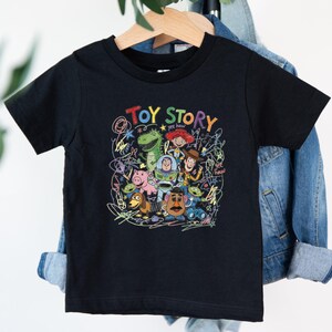 May include: A black short-sleeved t-shirt featuring a colorful graphic print of Toy Story characters including Woody, Buzz Lightyear, Jessie, Rex, Hamm, and Mr. Potato Head, surrounded by scribbled line art and the text Toy Story and yee haw.